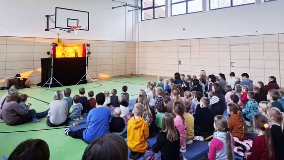 zeigt Aufführung mit zusehenden sitzenden Kindern und Erwachsenen