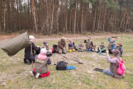 zeigt auf dem Boden sitzende Kindergruppe und Erwachsene im Gespräch
