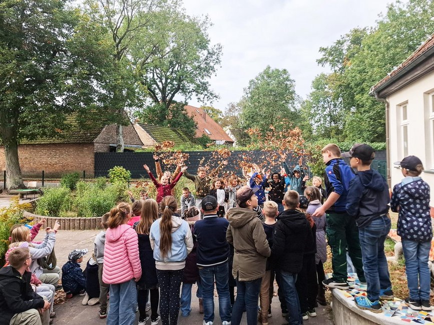 zeigt Kindergruppe, die mit Laub einen Blätterregen imitiert und weitere Kinder, die zuschauen